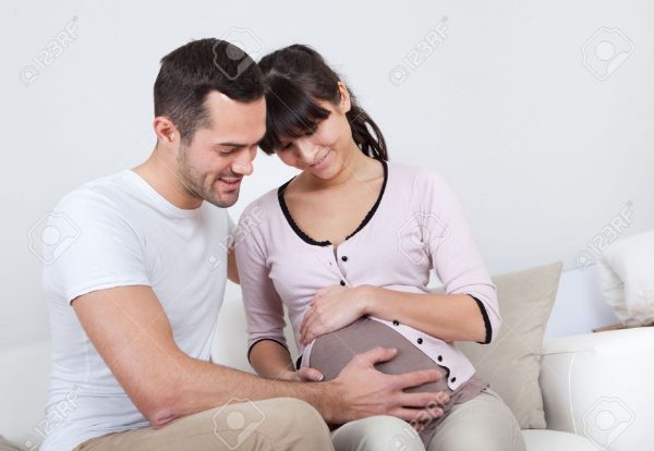 13020618-portrait-of-happy-pregnant-woman-and-her-husband-on-sofa-at-home-stock-photo